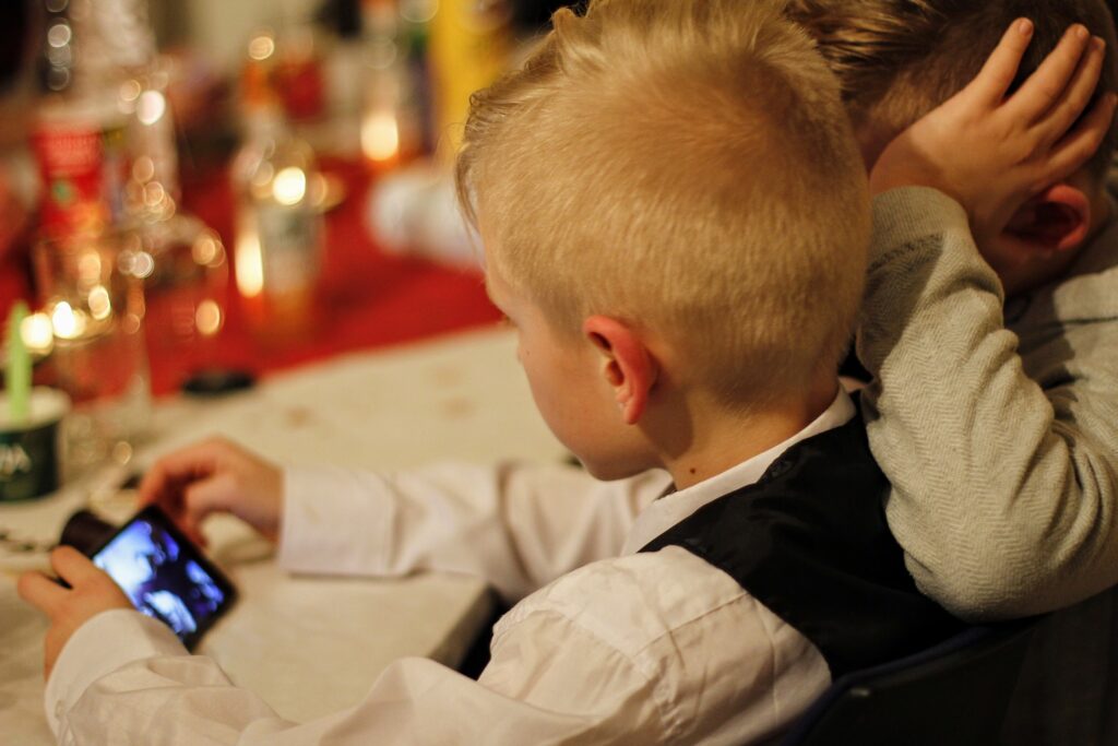 Deux garçons collés l'un à l'autre pour regarder l'écran d'un téléphone portable tenu par l'un des deux, en appui sur une table qui semble décorée pour une fête. L'écran diffuse une vidéo qu'il est impossible d'identifier