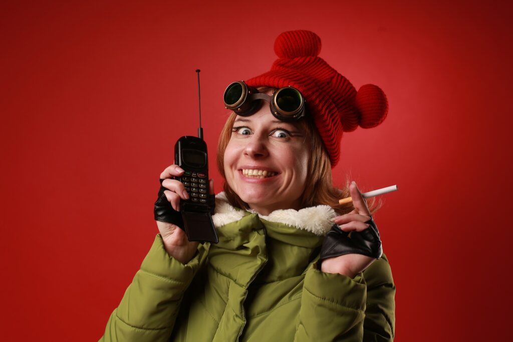 Photo d'une jeune femme rousse portant un bonnet à oreilles rouges, des lunettes d'aviateur, des mitaines en cuir et un anorak vert. Elle a un large sourire qui semble forcé et qui lui donne un air farfelu. Dans sa main droite, elle tient un téléphone portable à clapet et à antenne télescopique comme dans les années 90. Dans sa main droite, une cigarette non encore allumée.