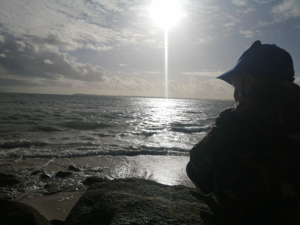 MMCC - paysage plage lorient mer rocher enfant casquette soleil contre jour nuages Photo d'un petit garçon de dos assis sur un rocher et regardant la mer sous le soleil couchant