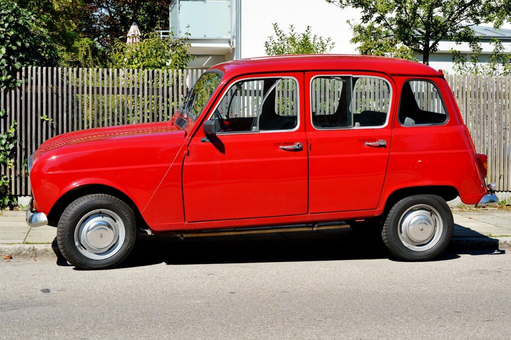 Photo d'une Renault 4L rouge vif garée sur un trottoir dans ce qui semble être un lotissement périurbain. Présence de barrières en bois, arbres et pavillon blanc en arrière plan.